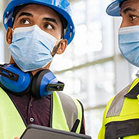 Image of 2 men wearing hard hats and blue surgical face coverings on a construction site