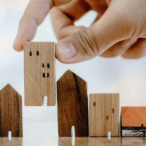 Image of a hand inserting a small wooden toy house, into a gap in a row of other toy wooden houses. 