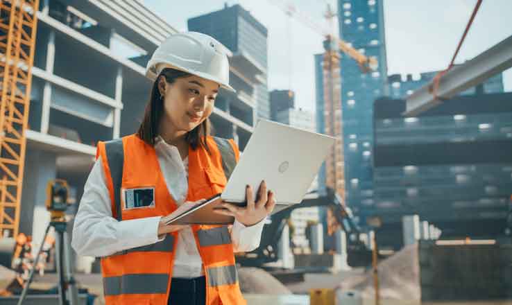 Woman on a building site looking at a computer, wearing an orange florescent jacket and wearing a hard helmet